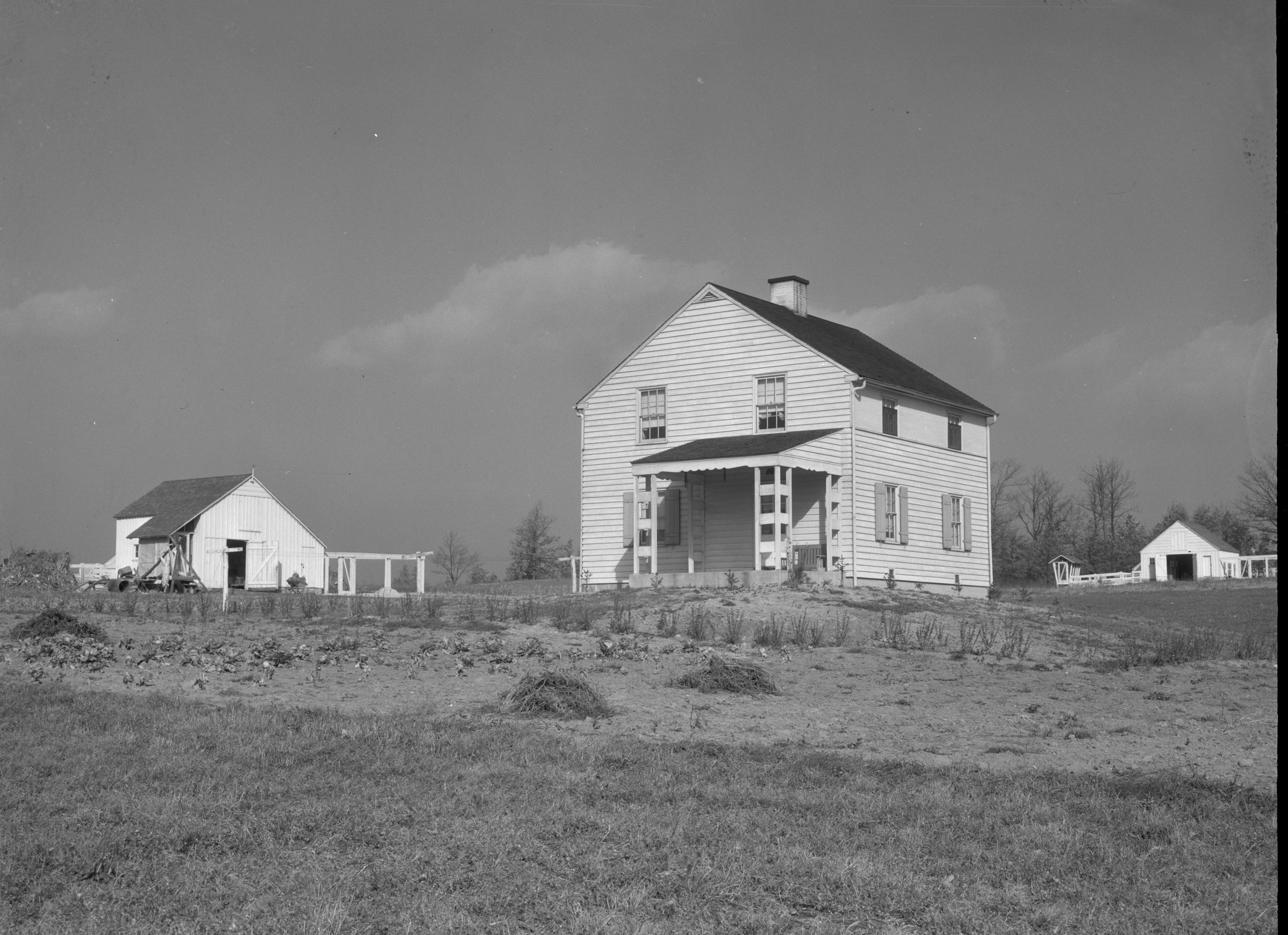 A white two-storey frame house with outbuildings on open fields under a clouded sky, Arthurdale, 1936