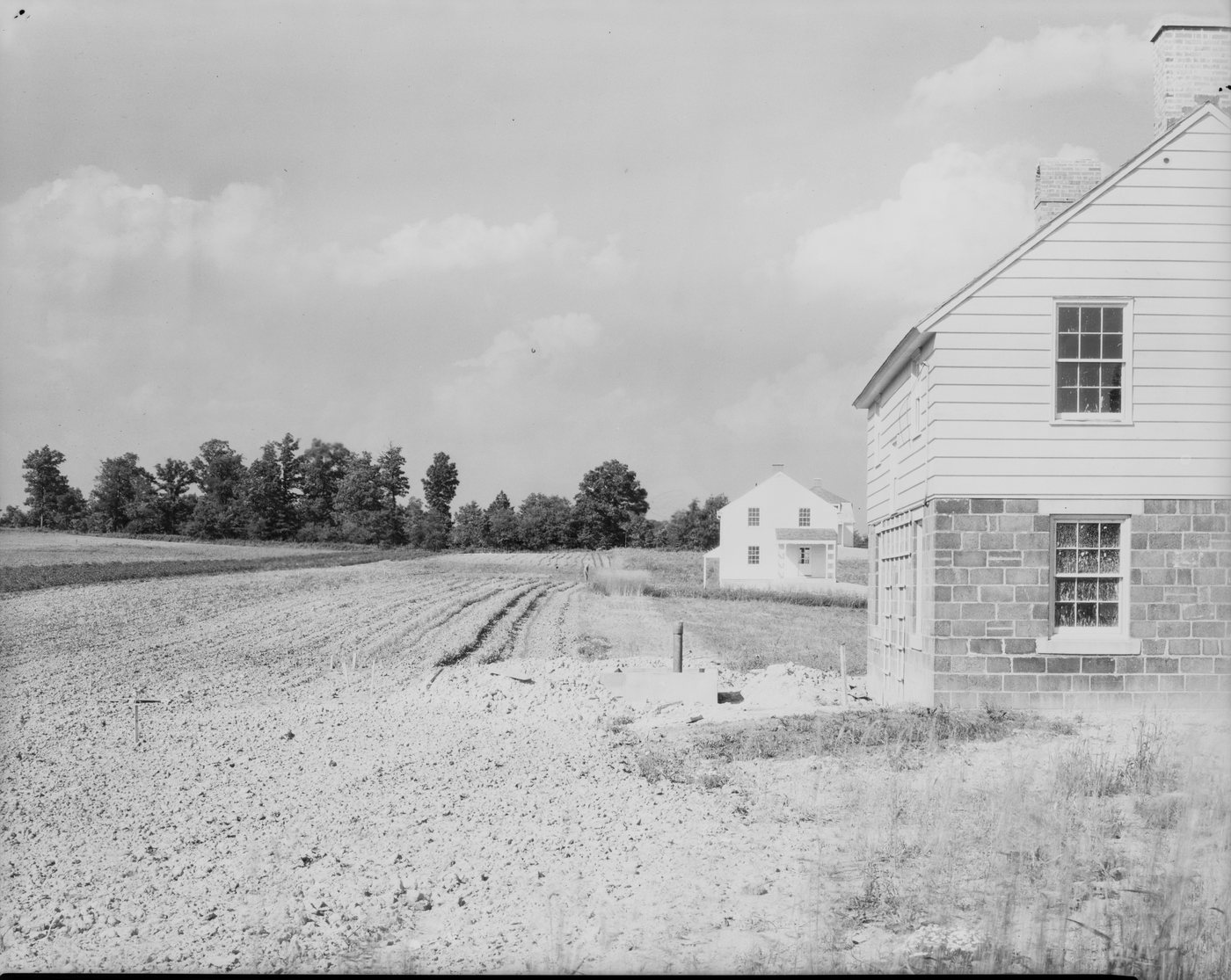 A cultivated field beside the corner of a stone-and-frame house, another house across the field