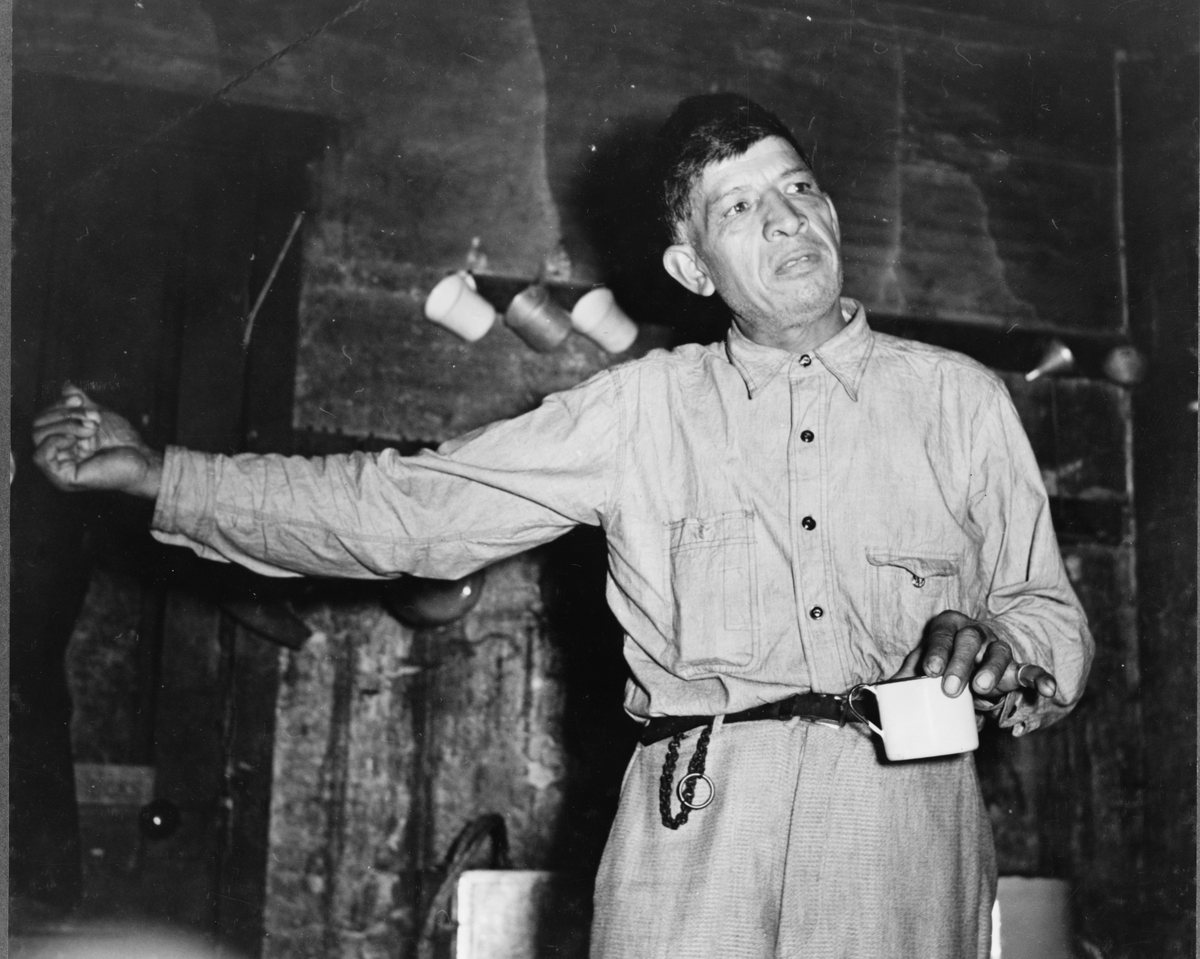 A man in work clothes with his arm outstretched, speaking, in front of a dark fireplace