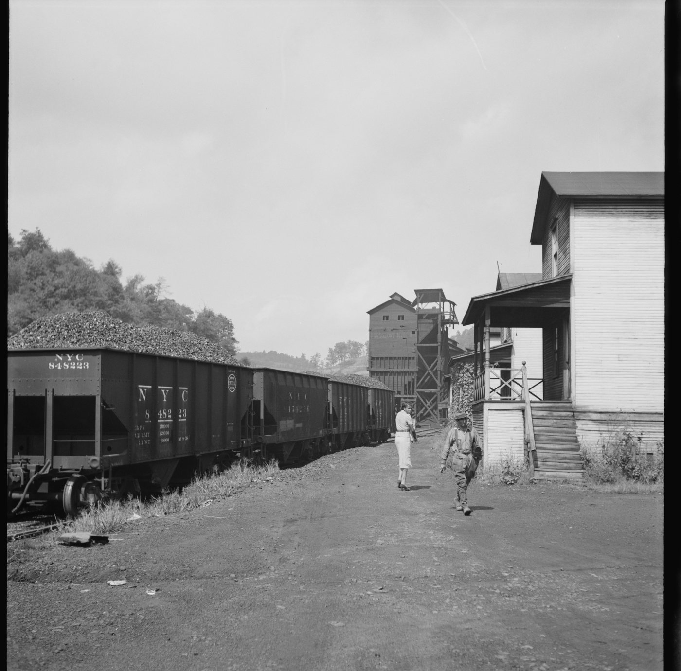 A dirt road between a line of coal cars and company houses, a tipple beyond, two people walking away