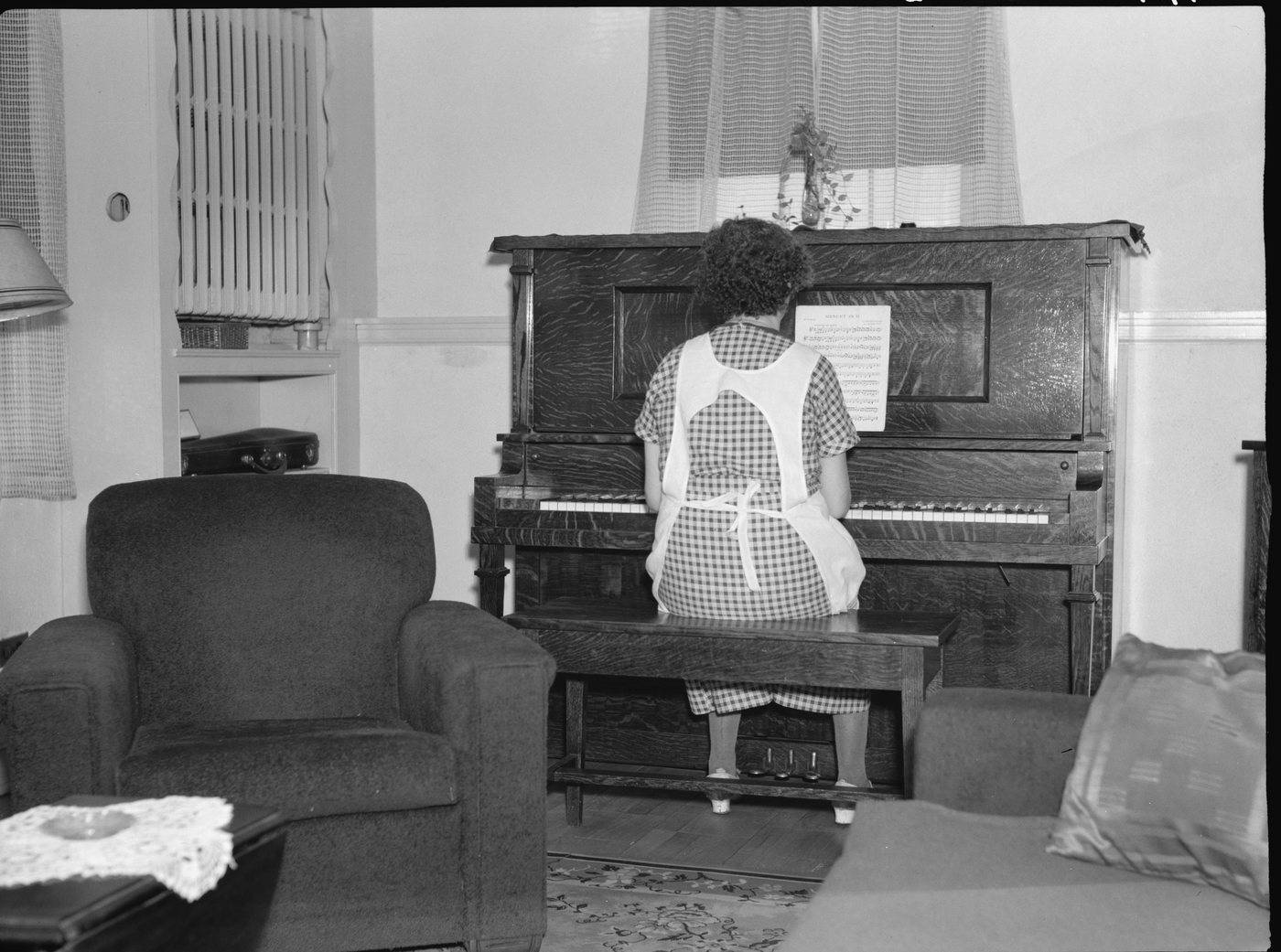 A woman in an apron playing an upright piano in a furnished living room
