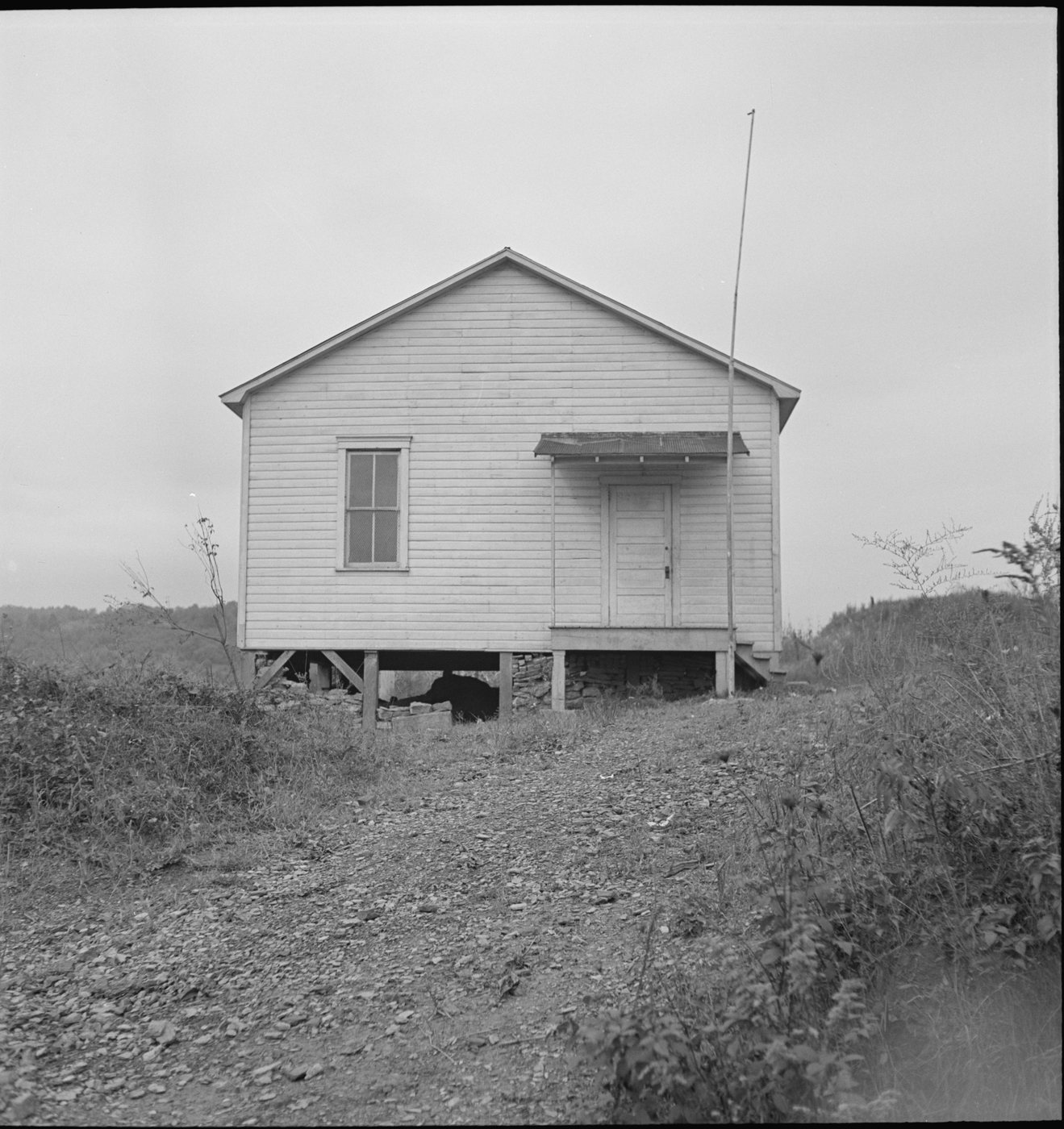 A small one-room frame school house on posts above a weedy slope