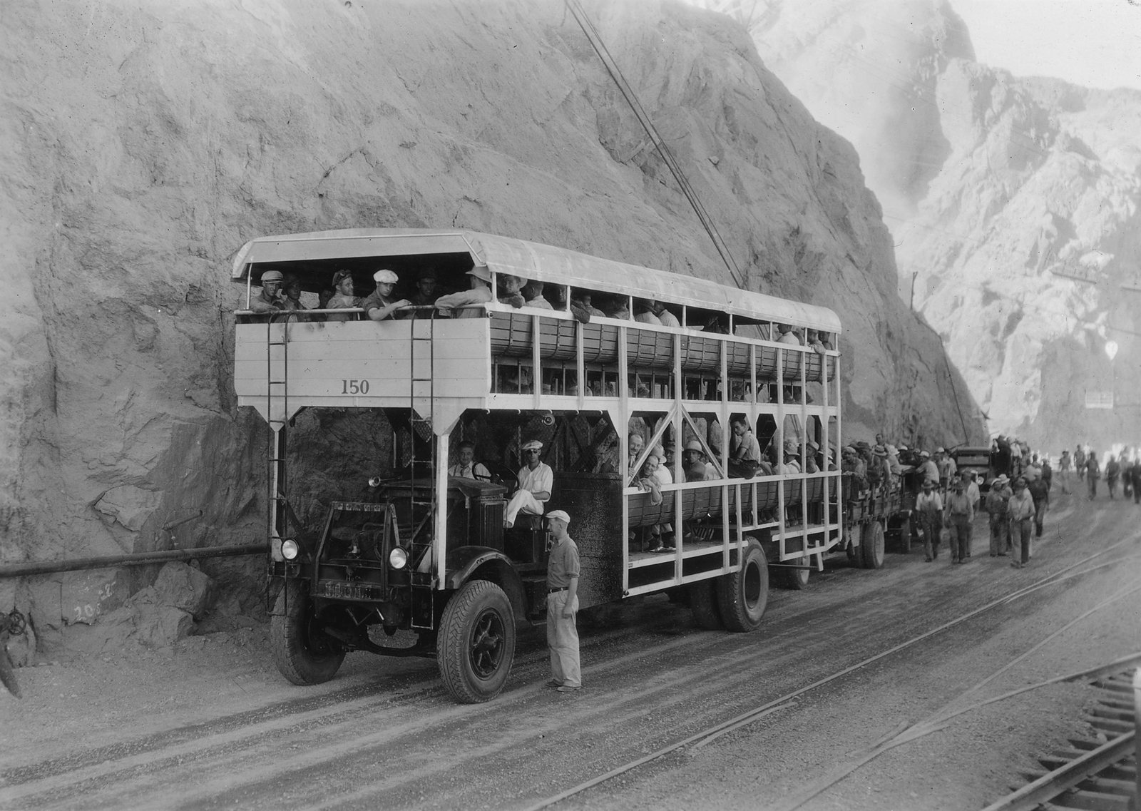 A double-decked open bus packed with workmen on a road cut into a canyon wall