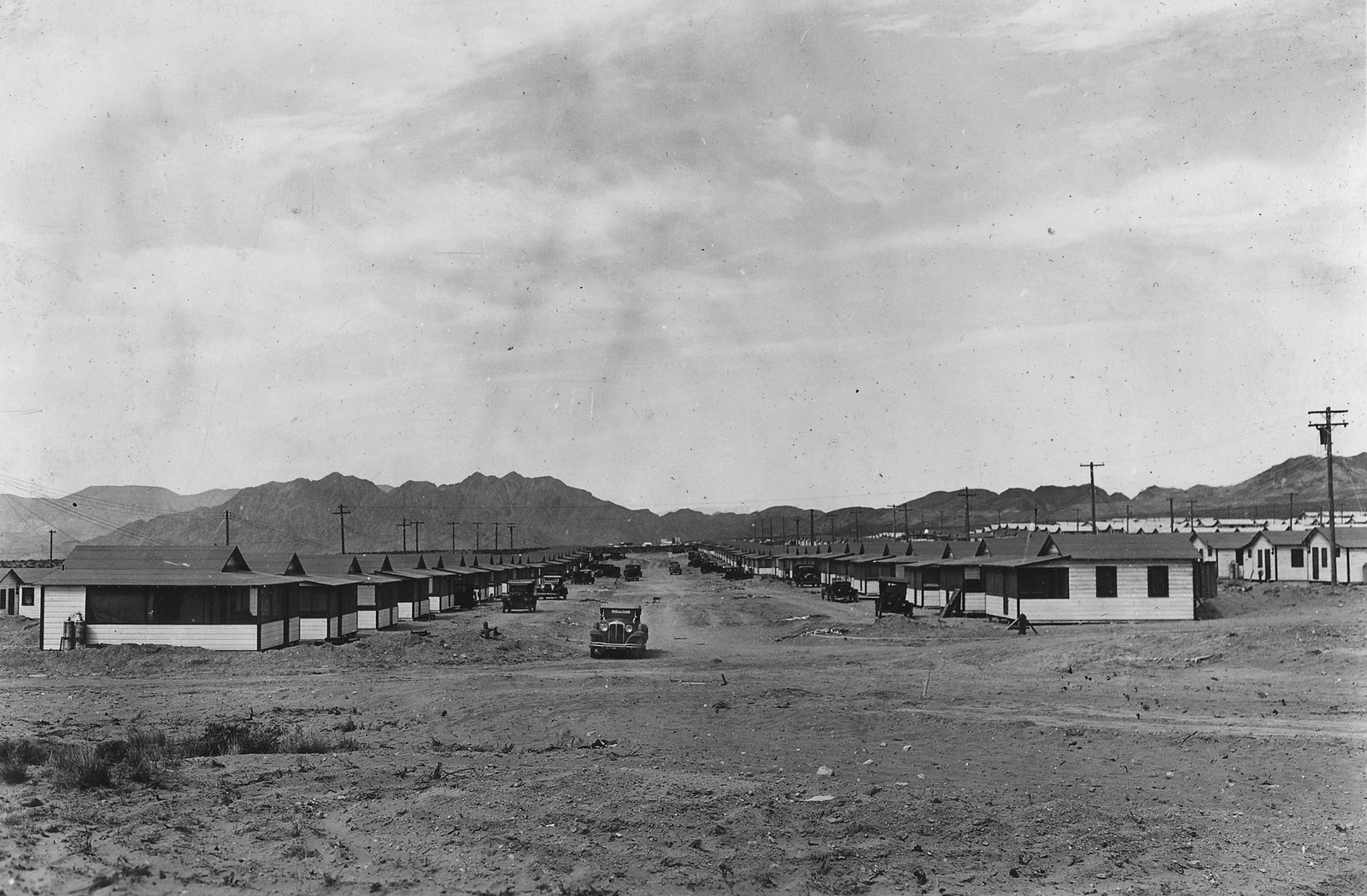 Two long rows of identical single-storey cottages along a bare dirt street, a parked car between them