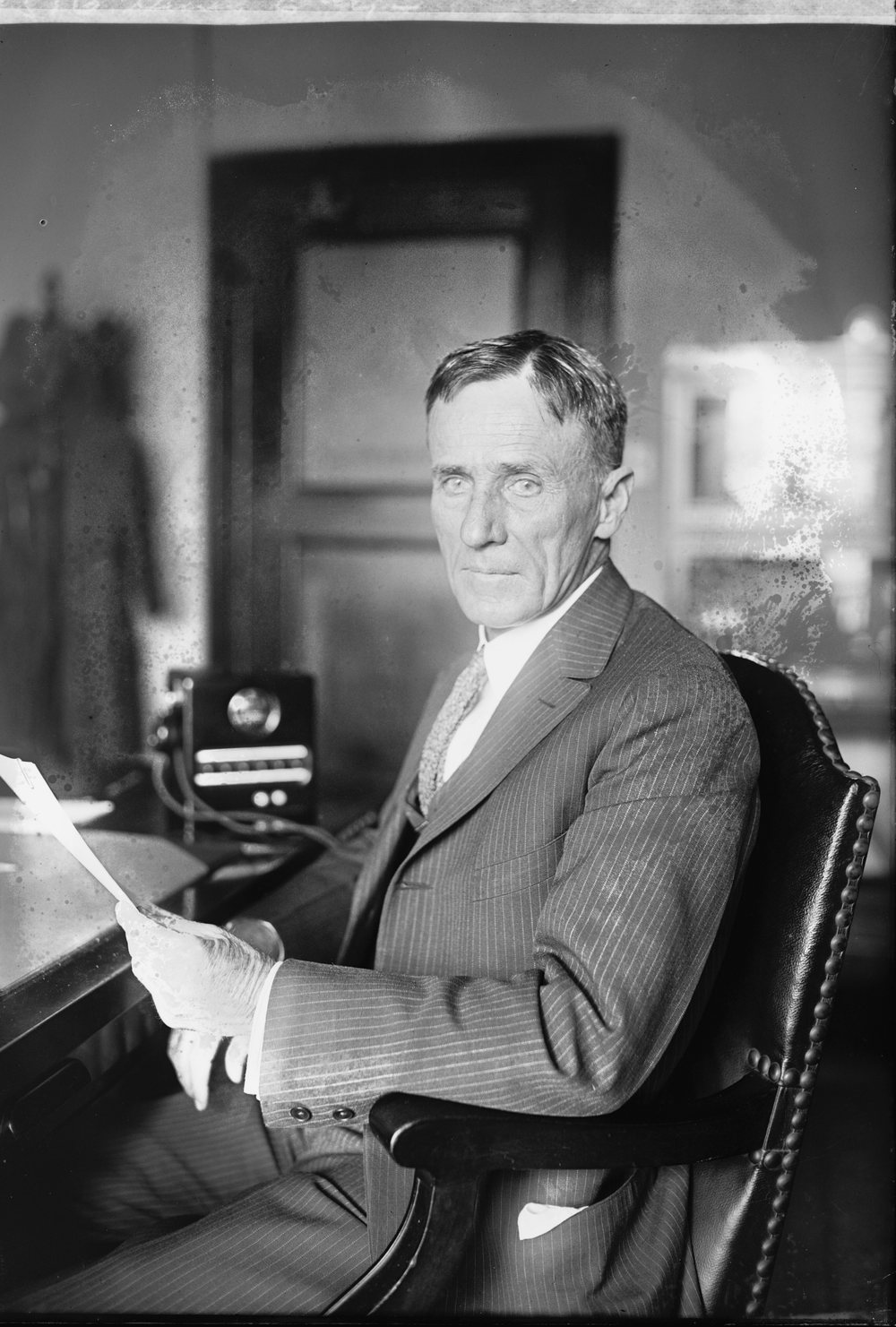 A grey-haired man in a dark suit seated at a desk with papers, a telephone at his elbow