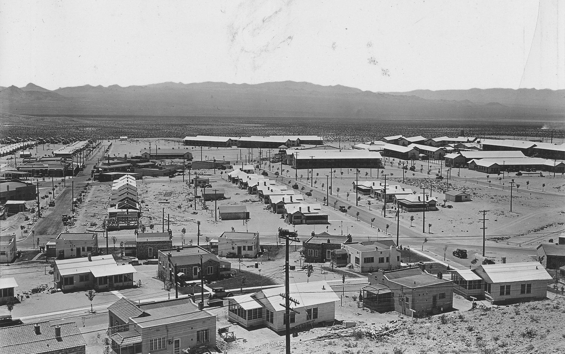 Rows of new single-storey houses and long low buildings spread across a desert flat below a hill, mountains on the horizon