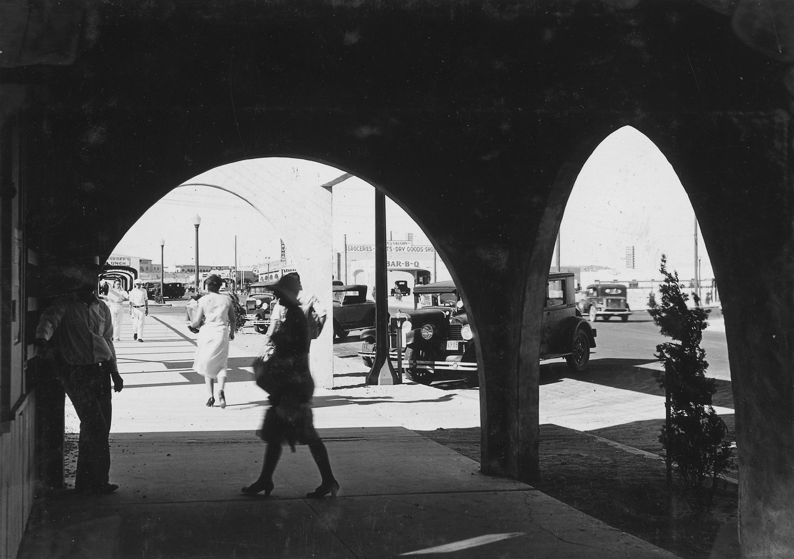 People walking past cars in sunlight, seen through the dark arches of an arcade
