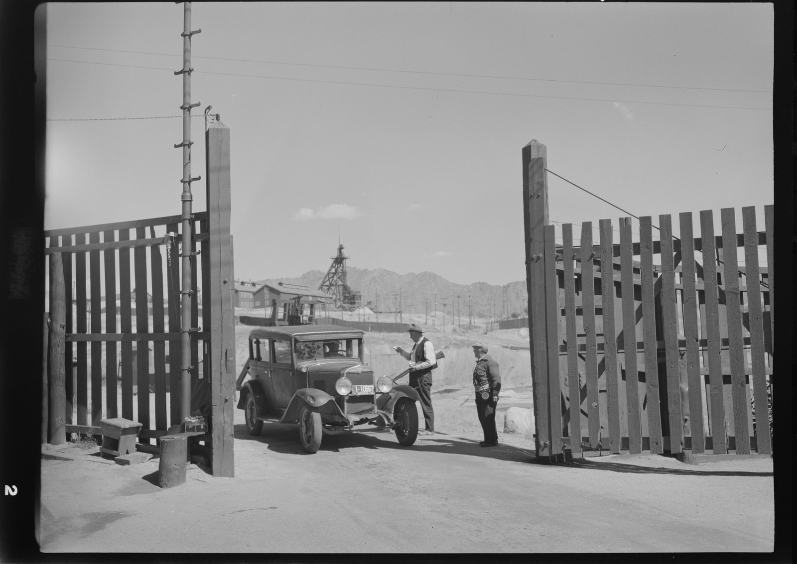 A car stopped at an open wooden gate while two guards speak to the driver, a headframe beyond