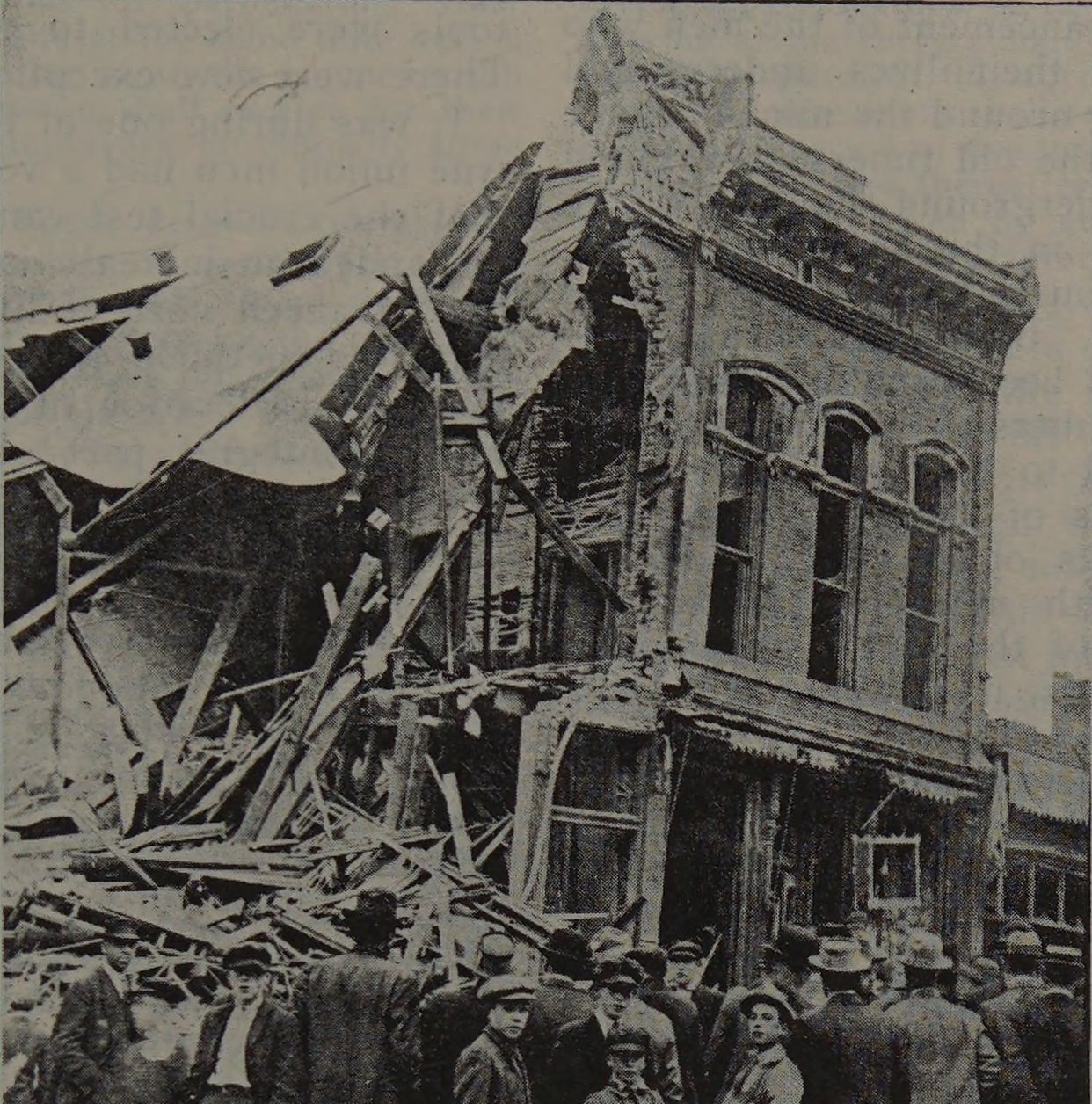A crowd in the street before the wrecked front of a brick building