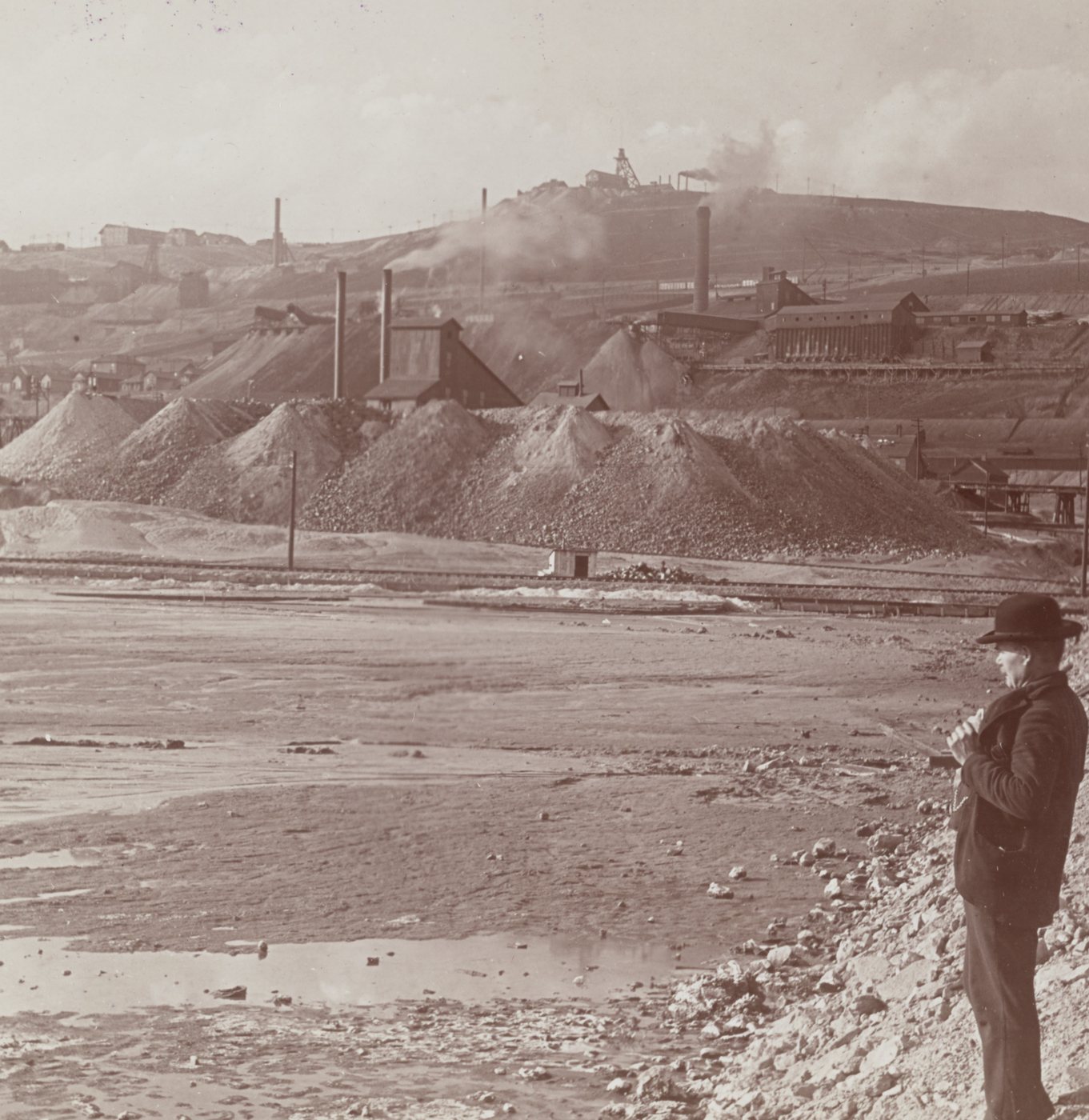A man in a bowler hat standing on bare ground before smoking mine works and waste heaps on a hill