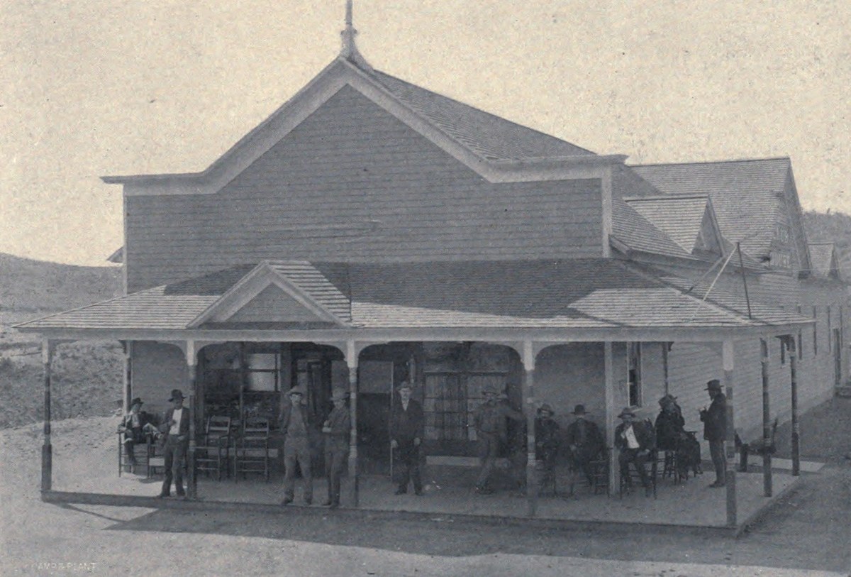 Men in hats on the deep veranda of a large gabled frame club house