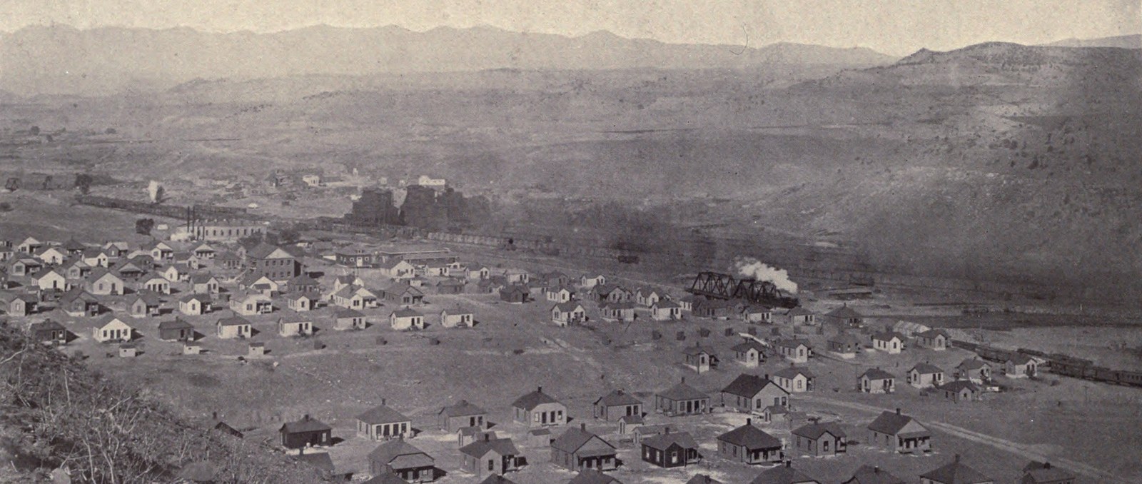 Rows of small company houses on a slope above a railway, washers and coke-oven smoke beyond, mountains on the horizon: Segundo in 1902