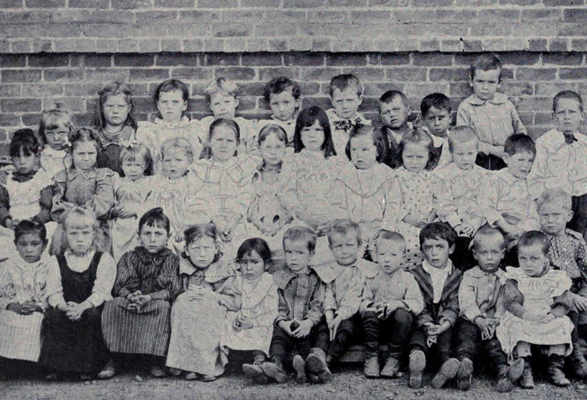 Several rows of small children in smocks and caps posed against a brick wall