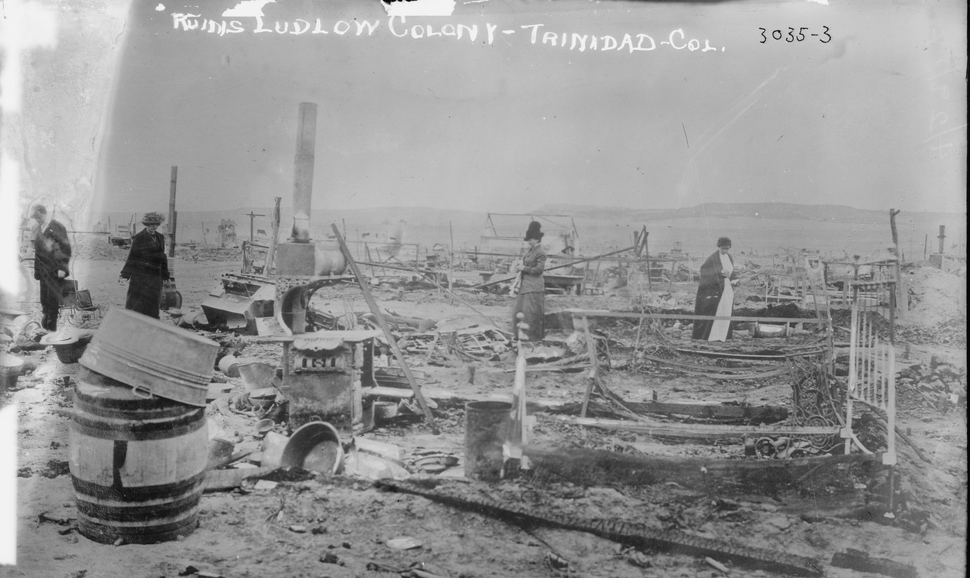 Women in long coats and hats walk among burnt bedsteads, a stove, a barrel and wreckage on bare ground
