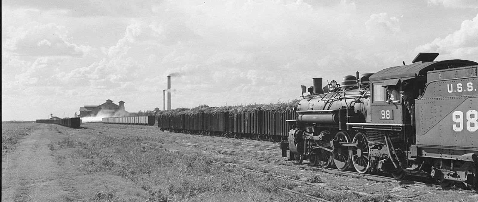 A steam locomotive lettered U.S.S.C. 98 pulling a long train of cane cars across flat fields, a sugar mill and its chimney on the horizon