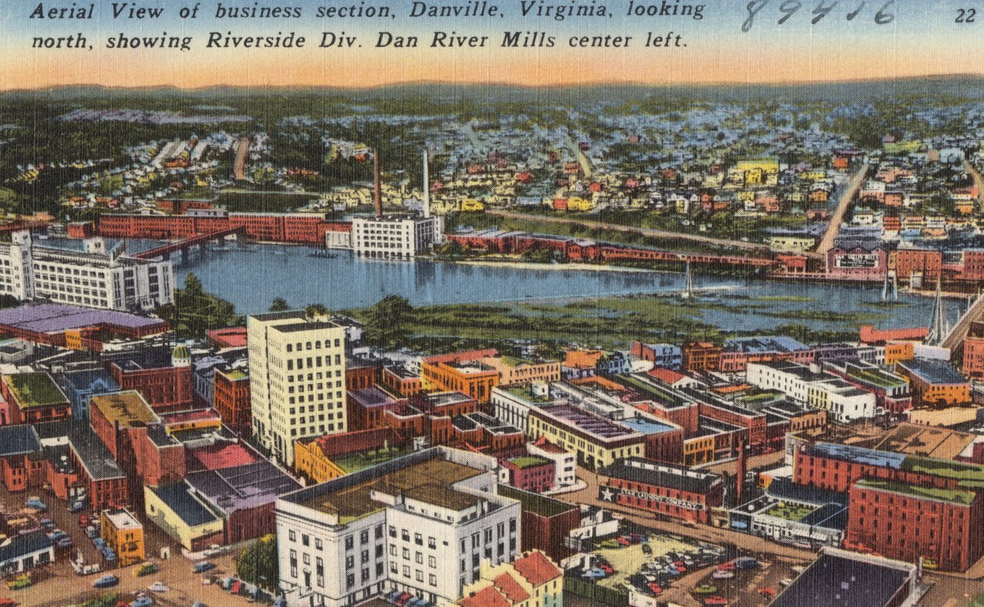 A coloured postcard aerial view of downtown Danville with a tall white building in front, the river across the middle and mill buildings on the far bank