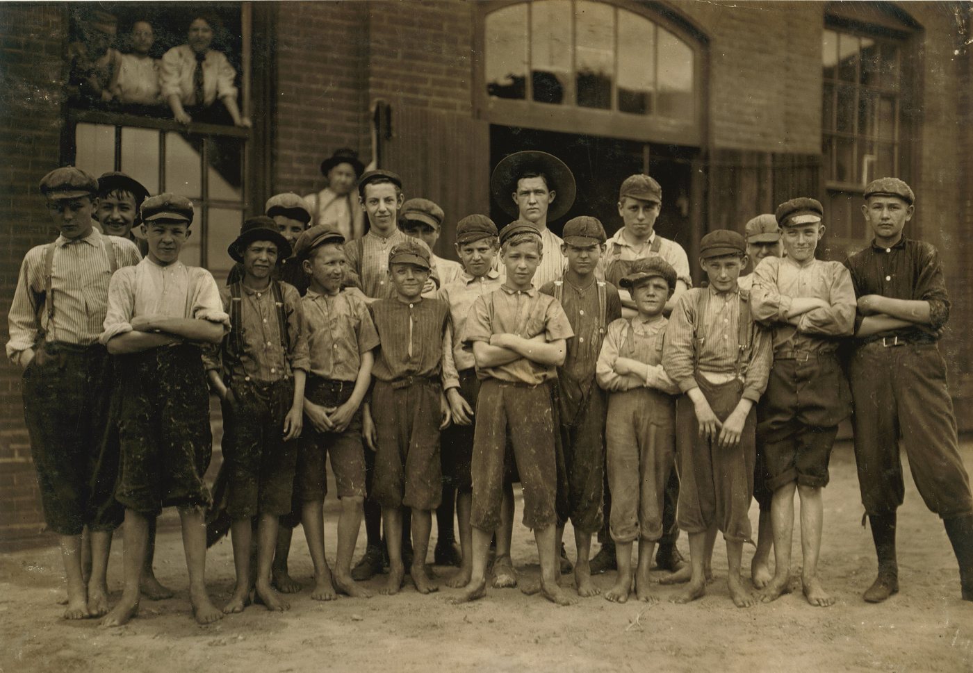 About twenty boys in caps and work clothes, many barefoot, standing in a group in front of a mill doorway