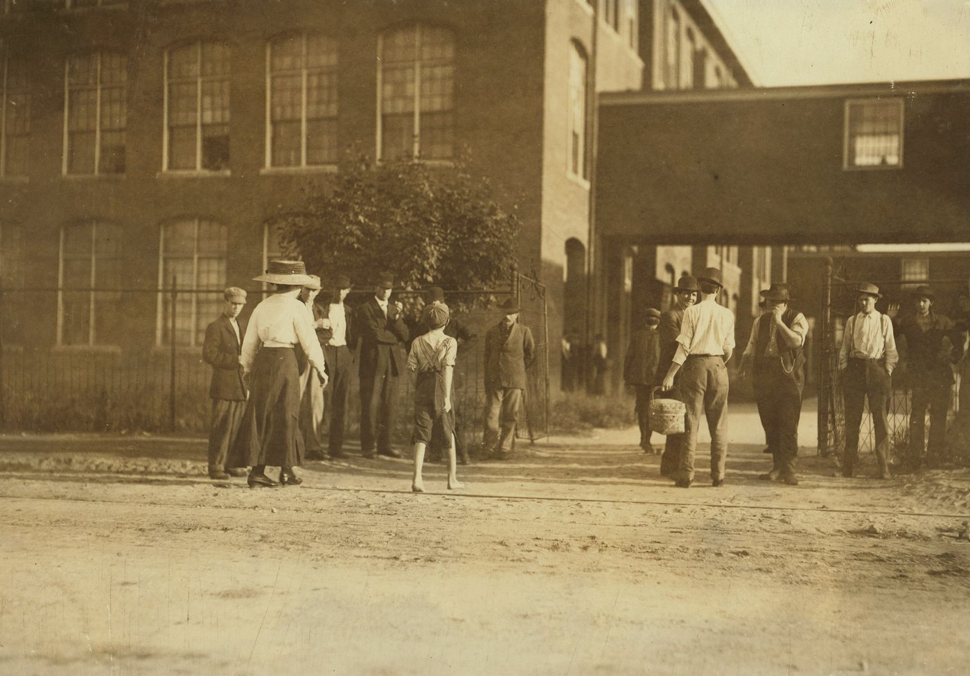 Men, women and a few children walking across a dirt yard toward a brick mill building in early morning light