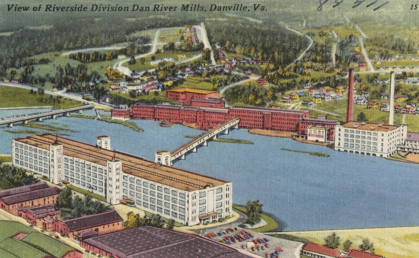 A coloured postcard aerial view of long red-brick and white mill buildings along a wide river, with a bridge and a tall chimney, and wooded hills behind