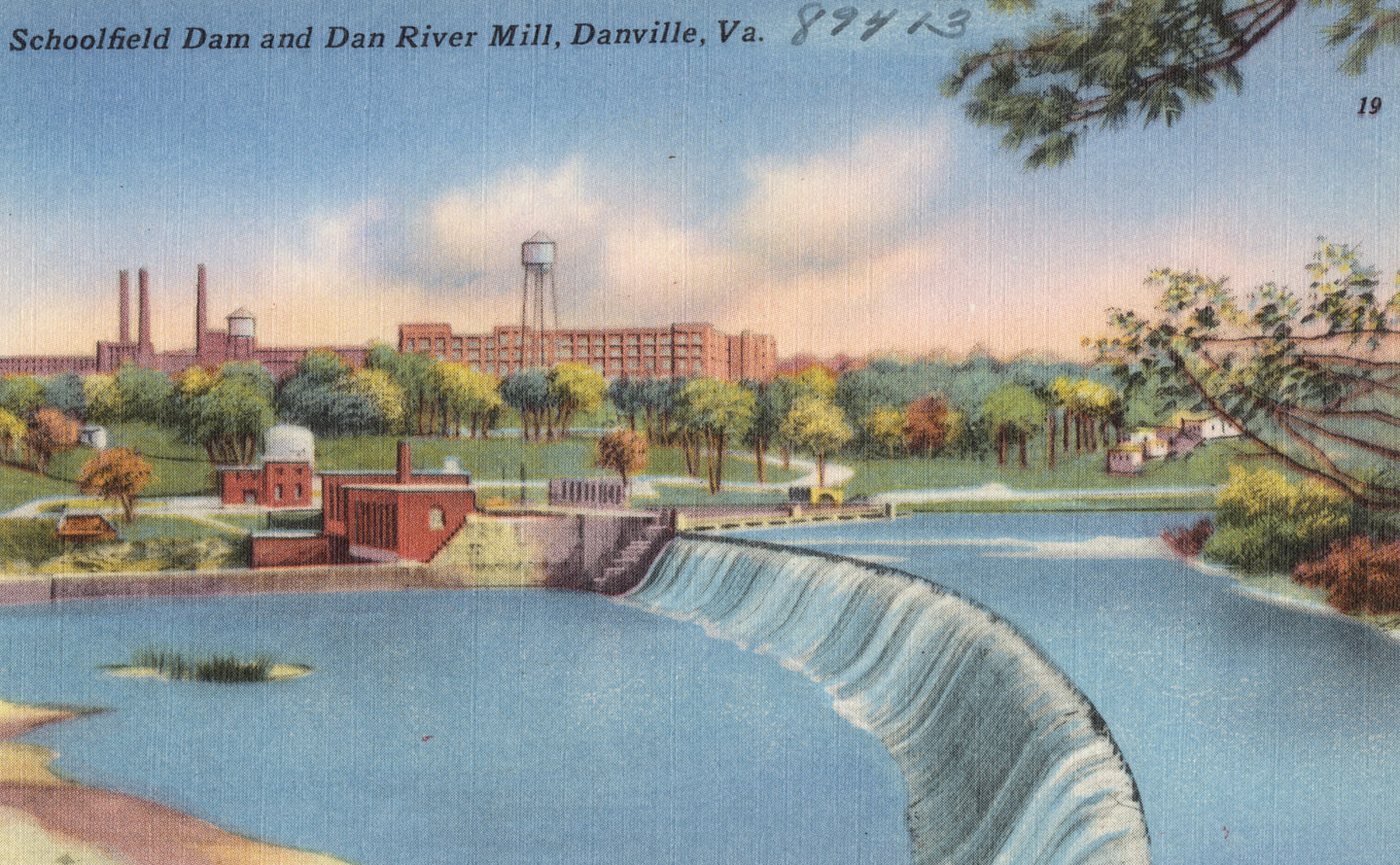 A coloured postcard of a curving dam with water pouring over it, and a long mill building with chimneys and a water tower beyond the trees