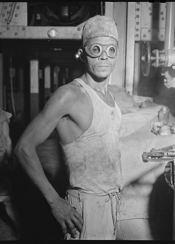 A man in a vest and cap with round goggles pushed up on his forehead, standing by machinery