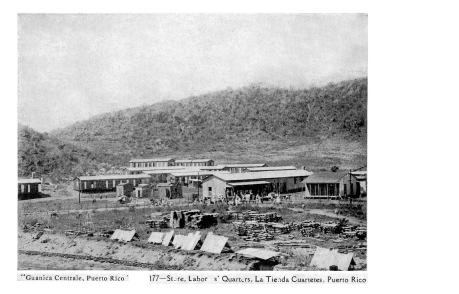 Long low buildings and rows of small houses on bare ground below a scrubby hill