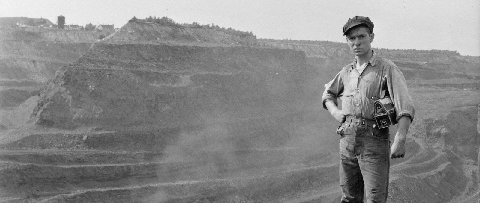 A young miner in a cap stands on broken rock at the edge of a vast terraced open pit, a camera case at his hip