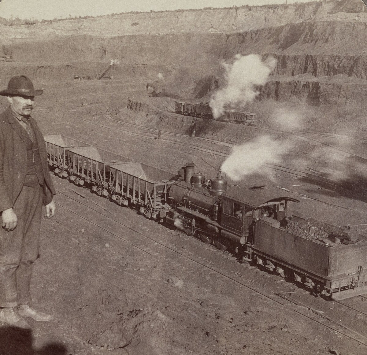 A man in a hat stands beside a steam locomotive hauling ore cars along the floor of an open pit, steam shovels working behind