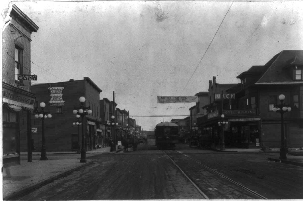 A paved main street with tram tracks, globe street lamps and brick shops, a streetcar in the distance