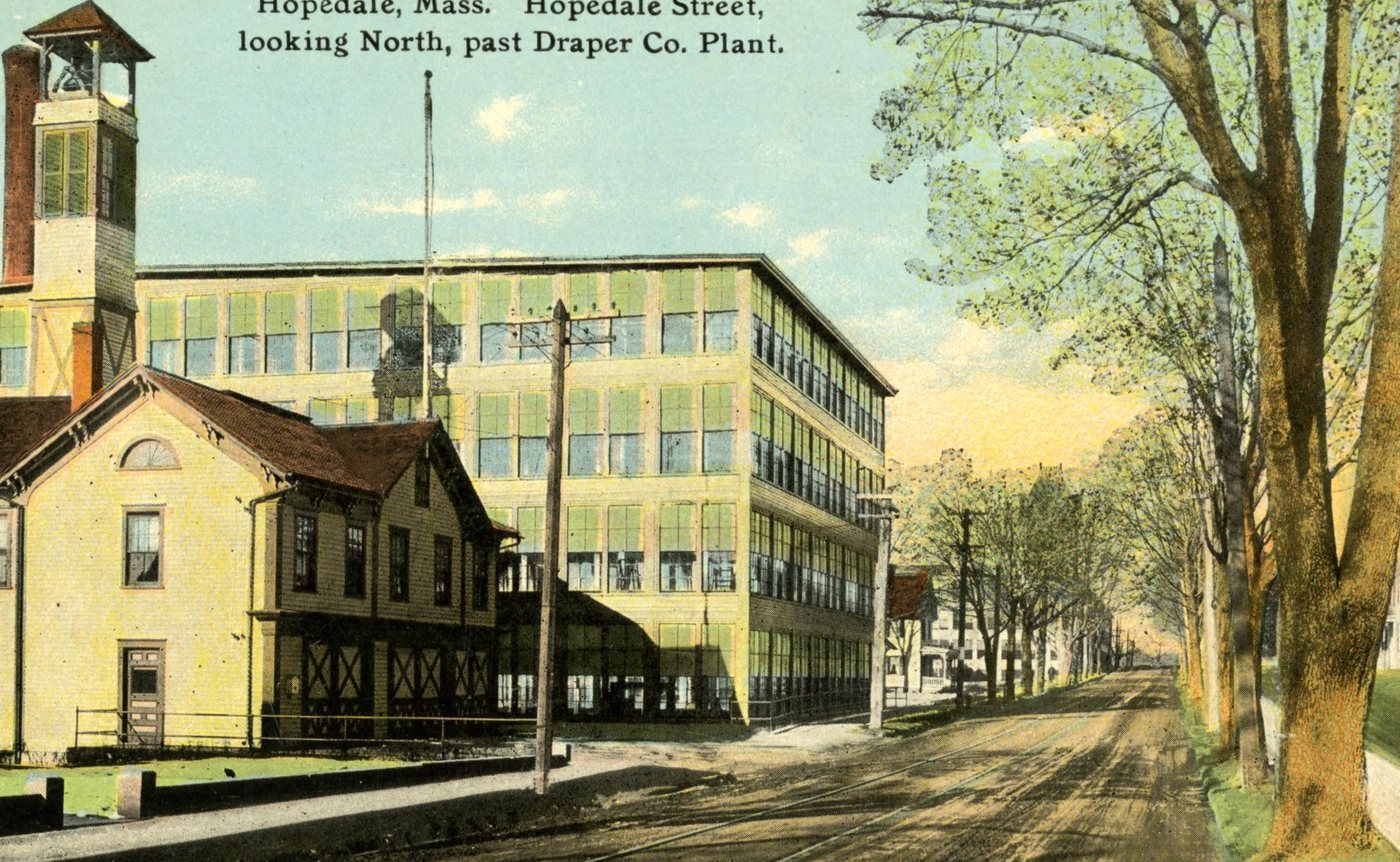 A hand-coloured postcard of Hopedale Street: a yellow wooden building with a bell tower in front of a long many-windowed mill, tracks running down a tree-lined street