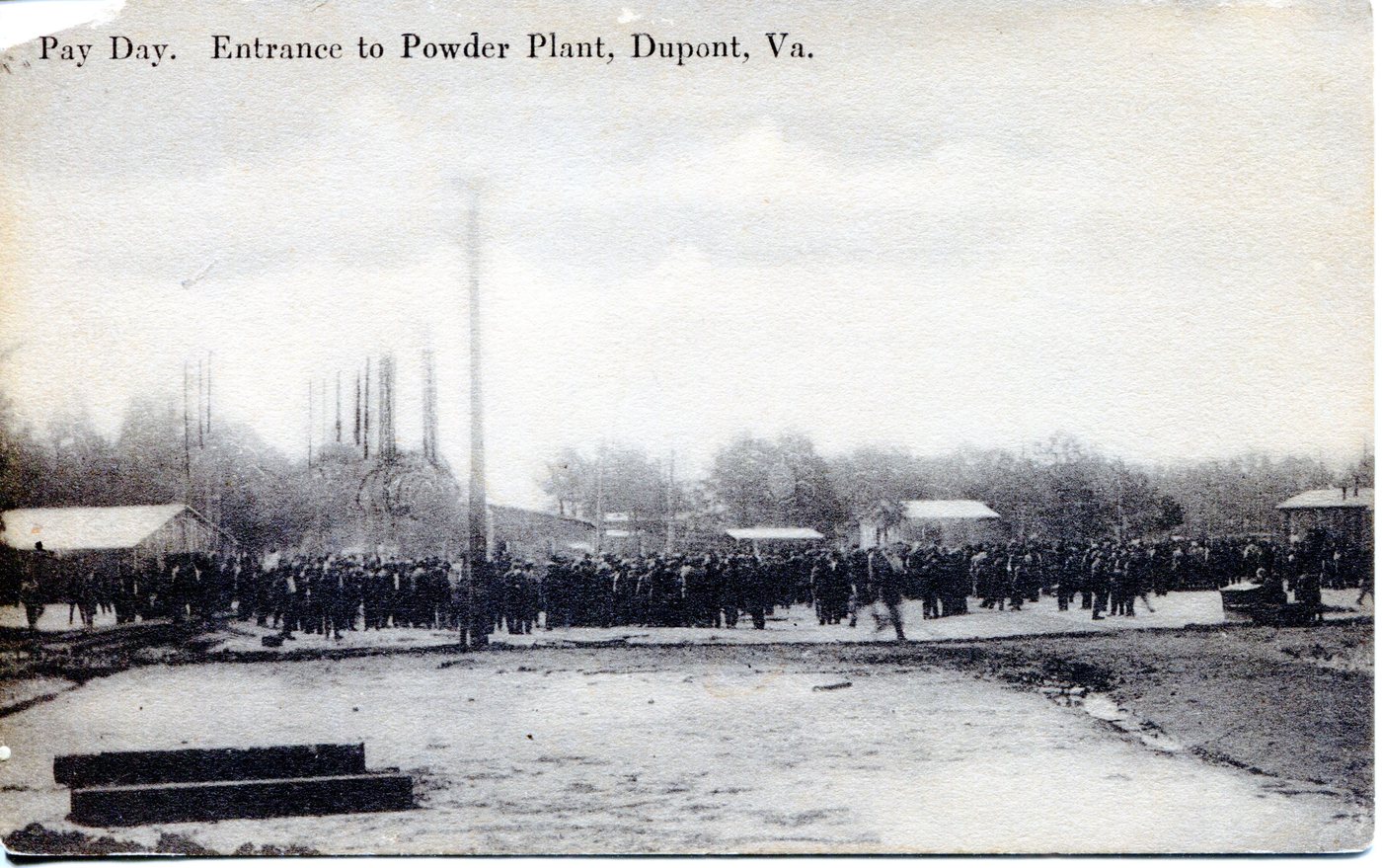 A large crowd of workers gathered at the gate of the powder plant, with stacks behind