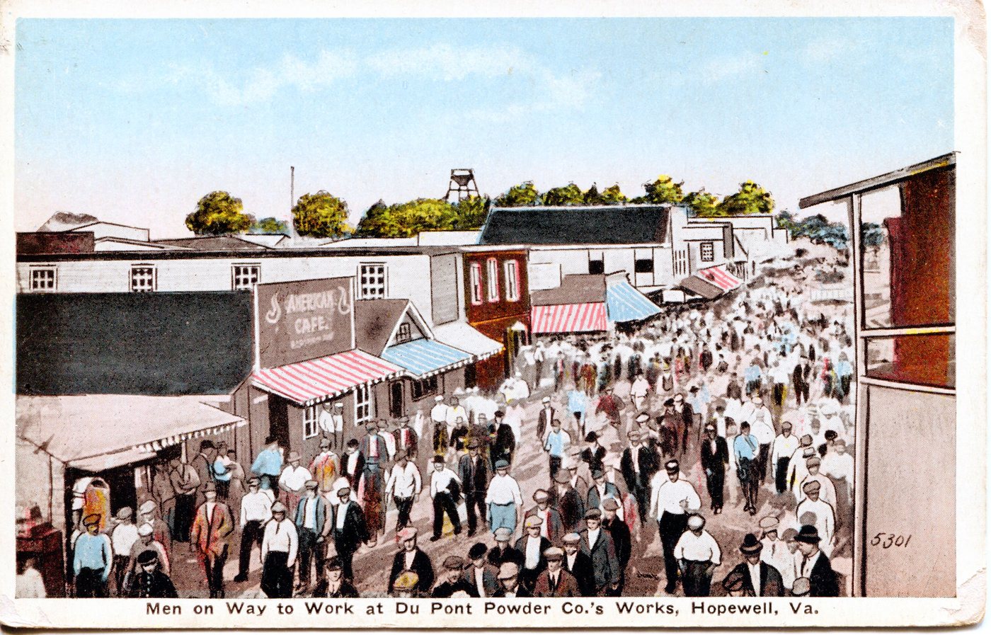 A hand-coloured postcard of a dense crowd of men in hats walking down a street of wooden shopfronts