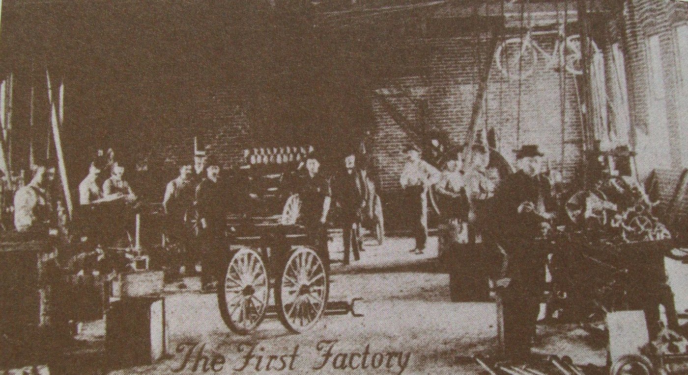 Men standing in a dim workshop around a carriage frame, with the words The First Factory in script