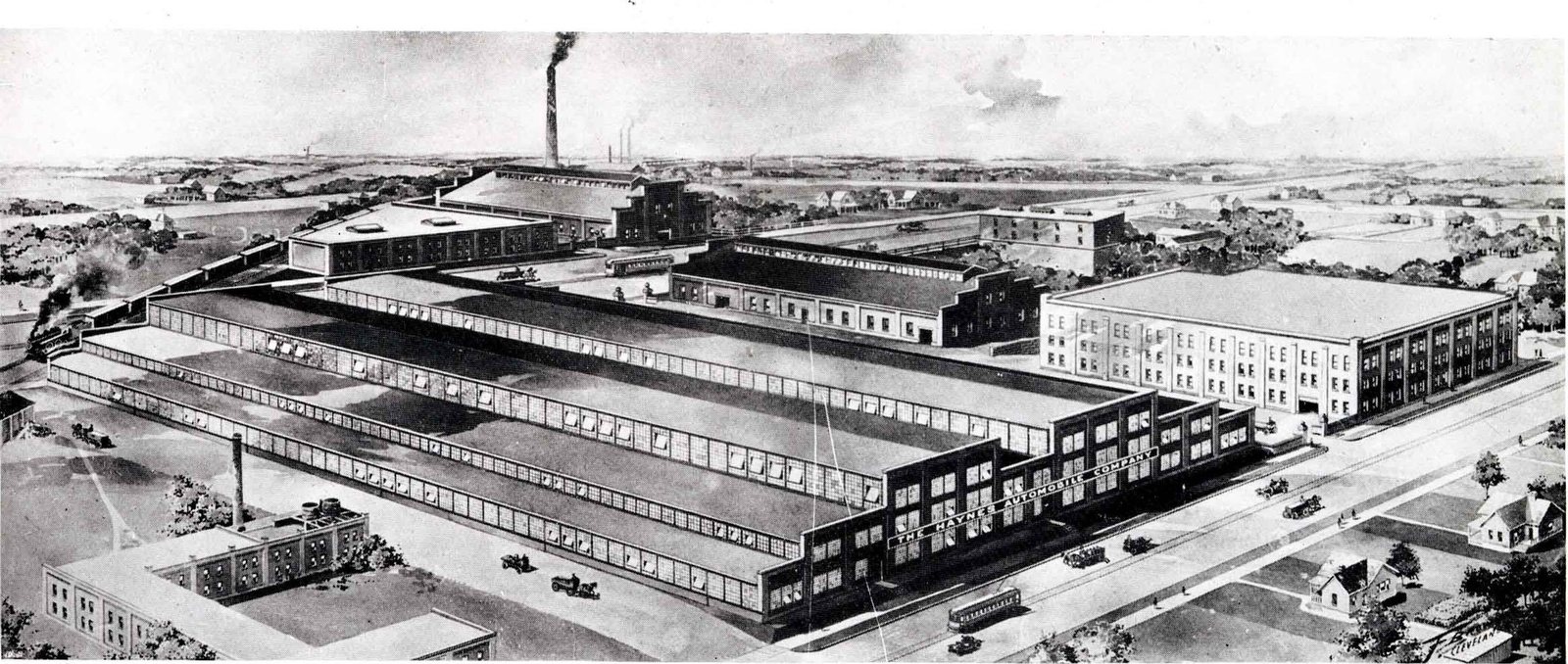 A bird's-eye view of a long low factory complex of sawtooth roofs and brick blocks, with a smokestack and houses beyond, Kokomo, 1916