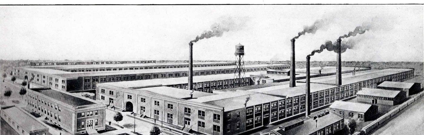 An aerial view of a large factory complex of long brick sheds with three tall smoking chimneys