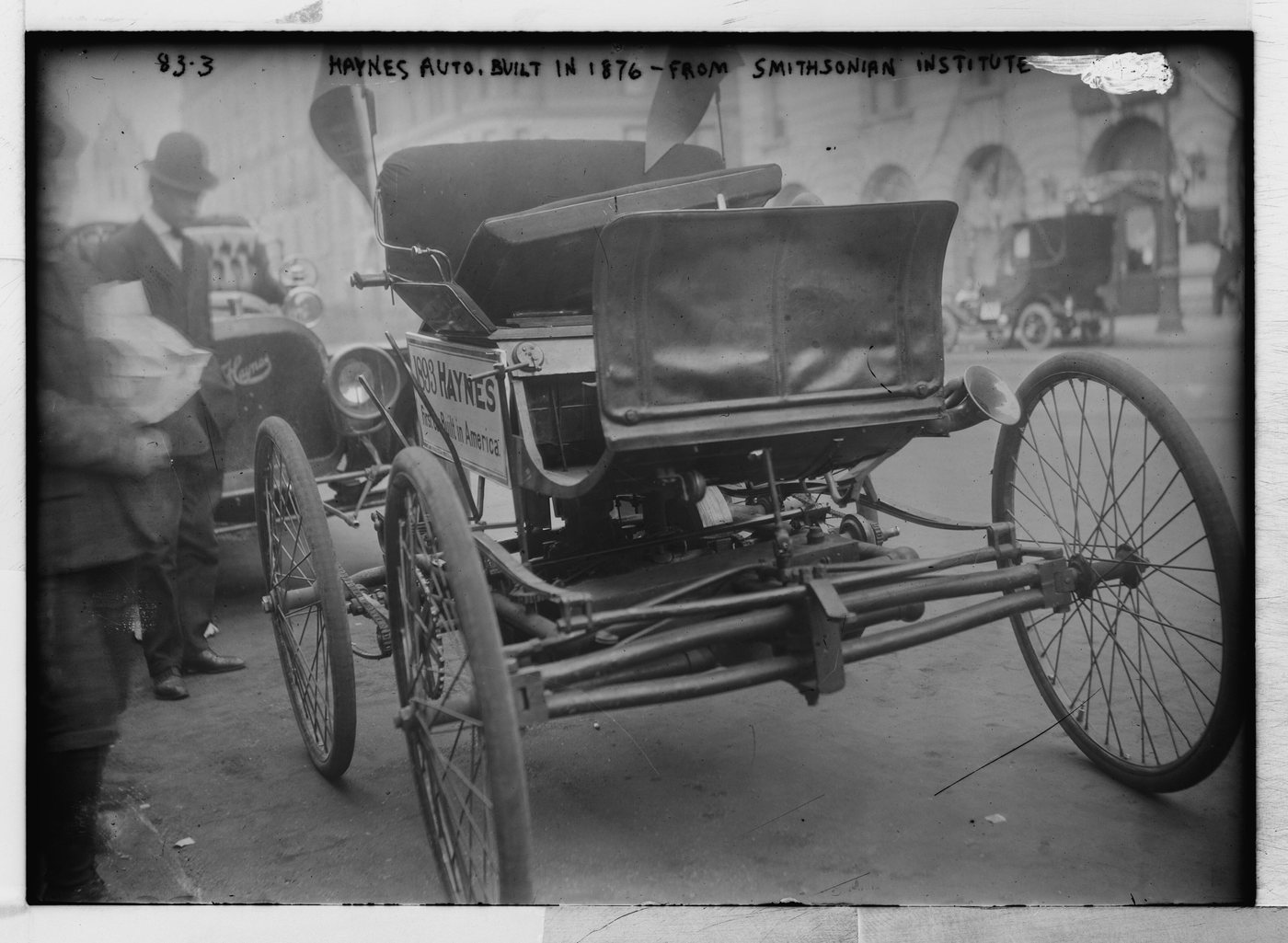 An early horseless carriage with high spindly wheels and a sign reading Haynes, parked on a city street with onlookers