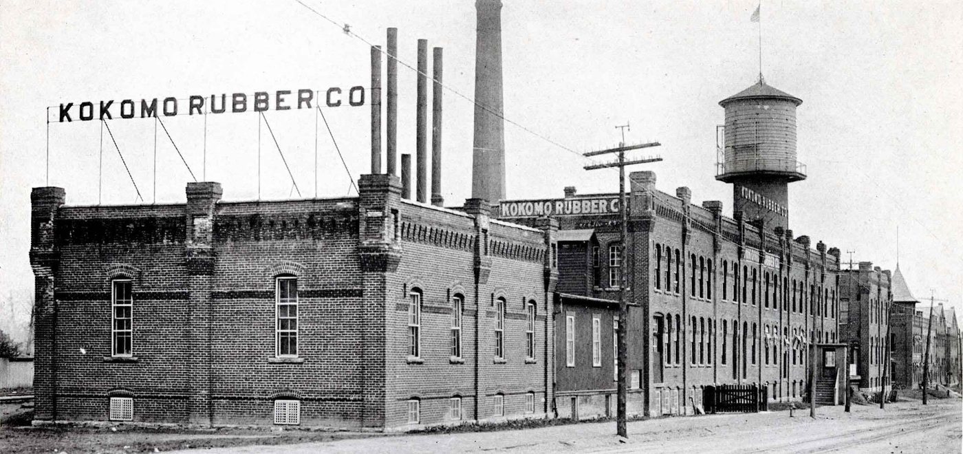 A long two-storey brick factory with the sign Kokomo Rubber Co on the roof, a water tower and four tall chimneys