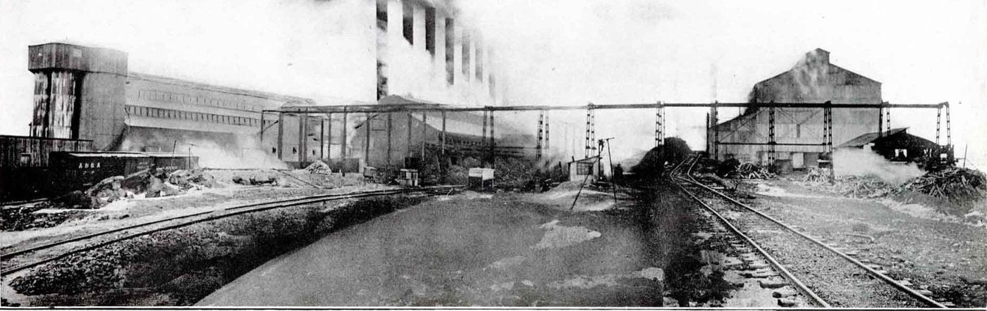 A long industrial works with smoking chimneys, a crane gantry and railway tracks in the foreground