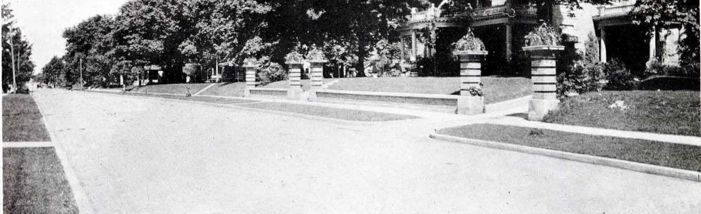 A wide tree-lined residential street with large porches and stone gateposts