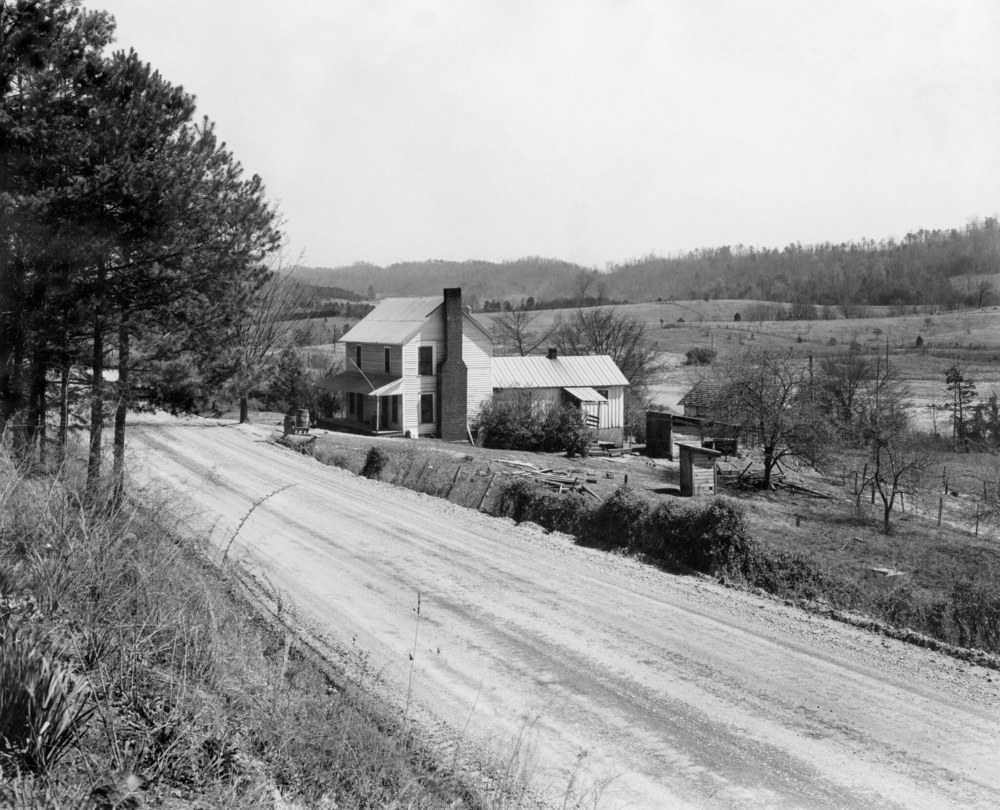 A white two-storey farmhouse beside a dirt road, with fields and wooded hills behind