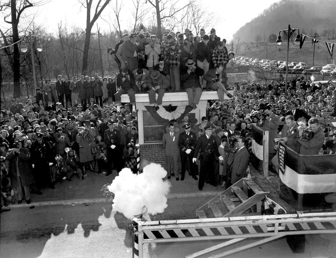 A large crowd in coats around a decorated platform at a gate, with a burst of white smoke rising in the foreground