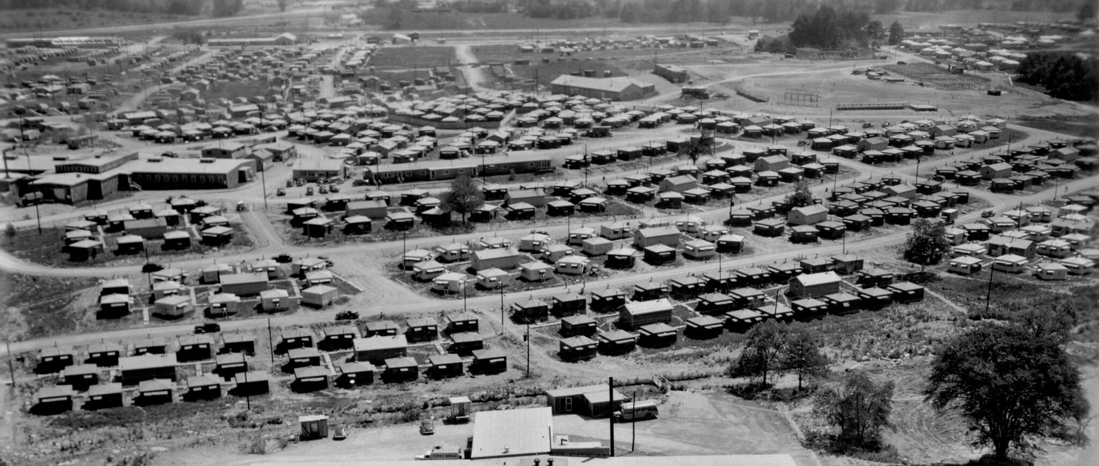 An aerial view of rows of small square plywood huts on bare ground, with a long low building and roads beyond, Oak Ridge, 1945