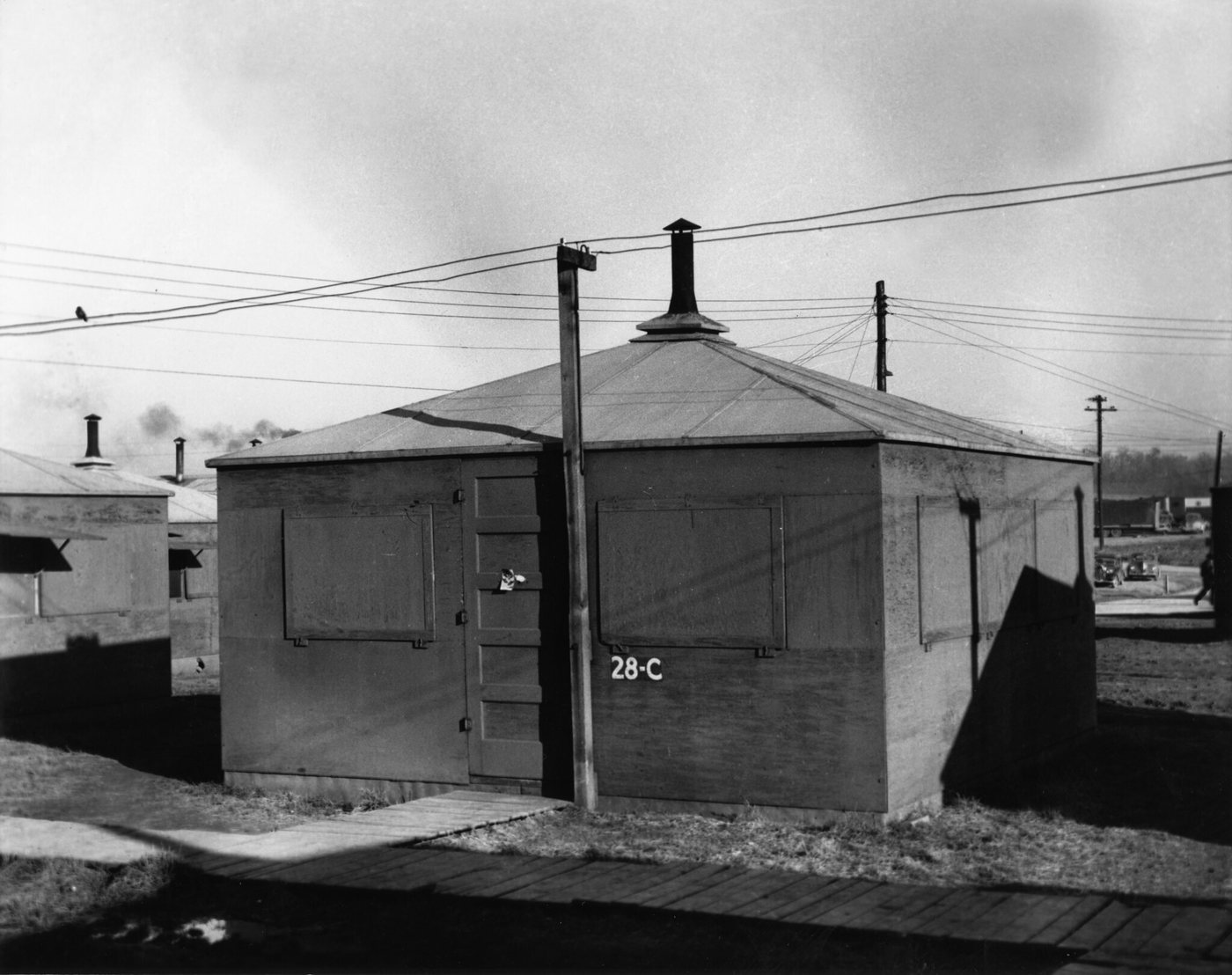 A square plywood hut with a pyramid roof, a stovepipe, closed wooden window shutters and the number 28-C painted on the side