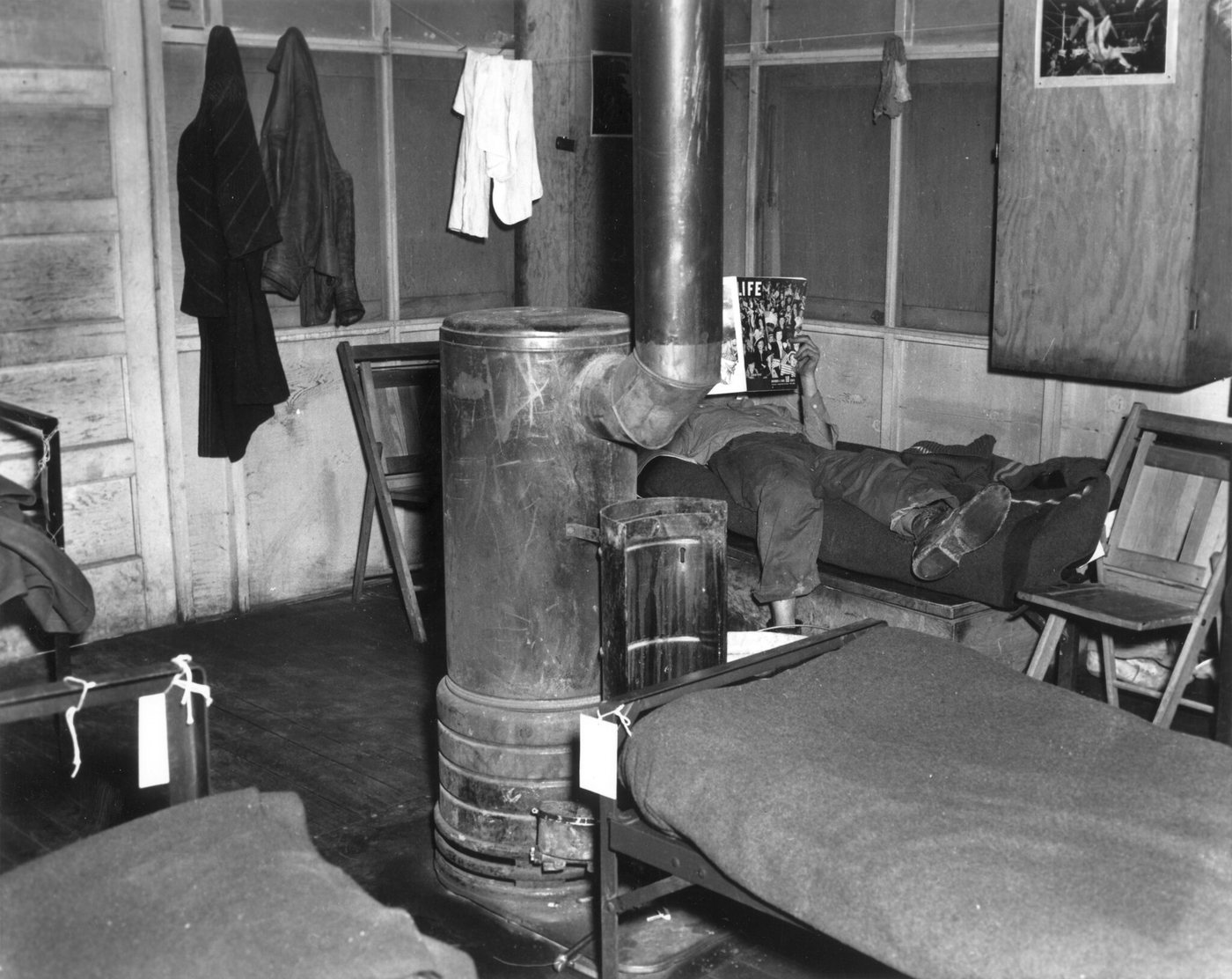 The inside of a hut: a cylindrical iron stove with a flue pipe, an army cot with a blanket, and clothes hanging on the wall