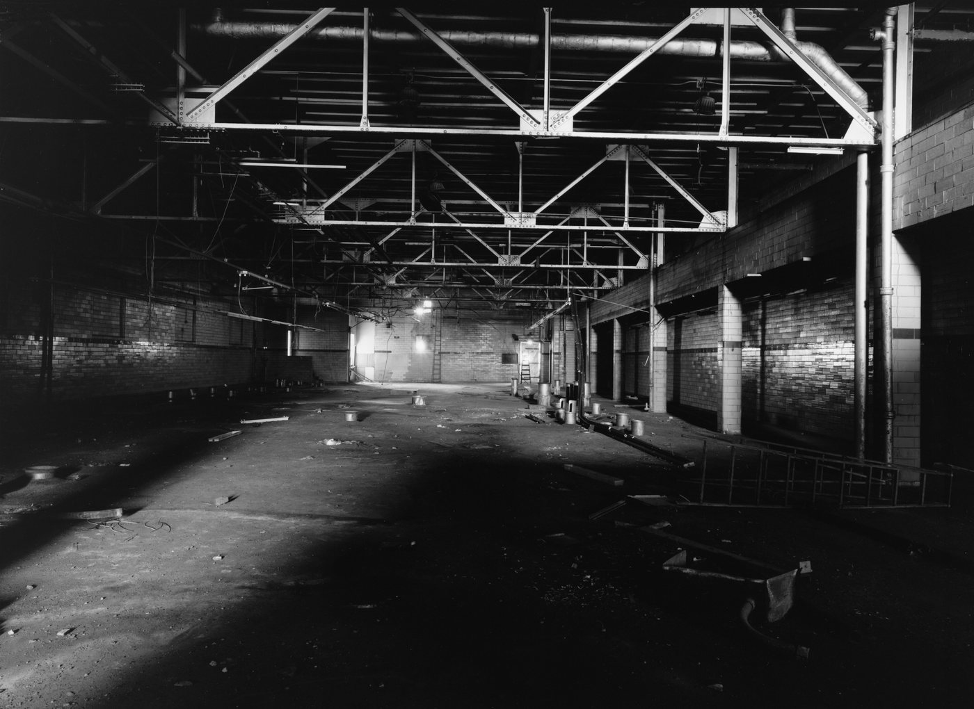 A long empty factory floor with steel roof trusses and a pool of light at the far end