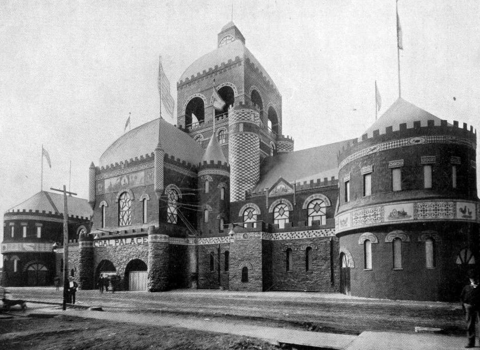 A large brick exhibition hall with round towers, battlements, domes and flags, seen from the street