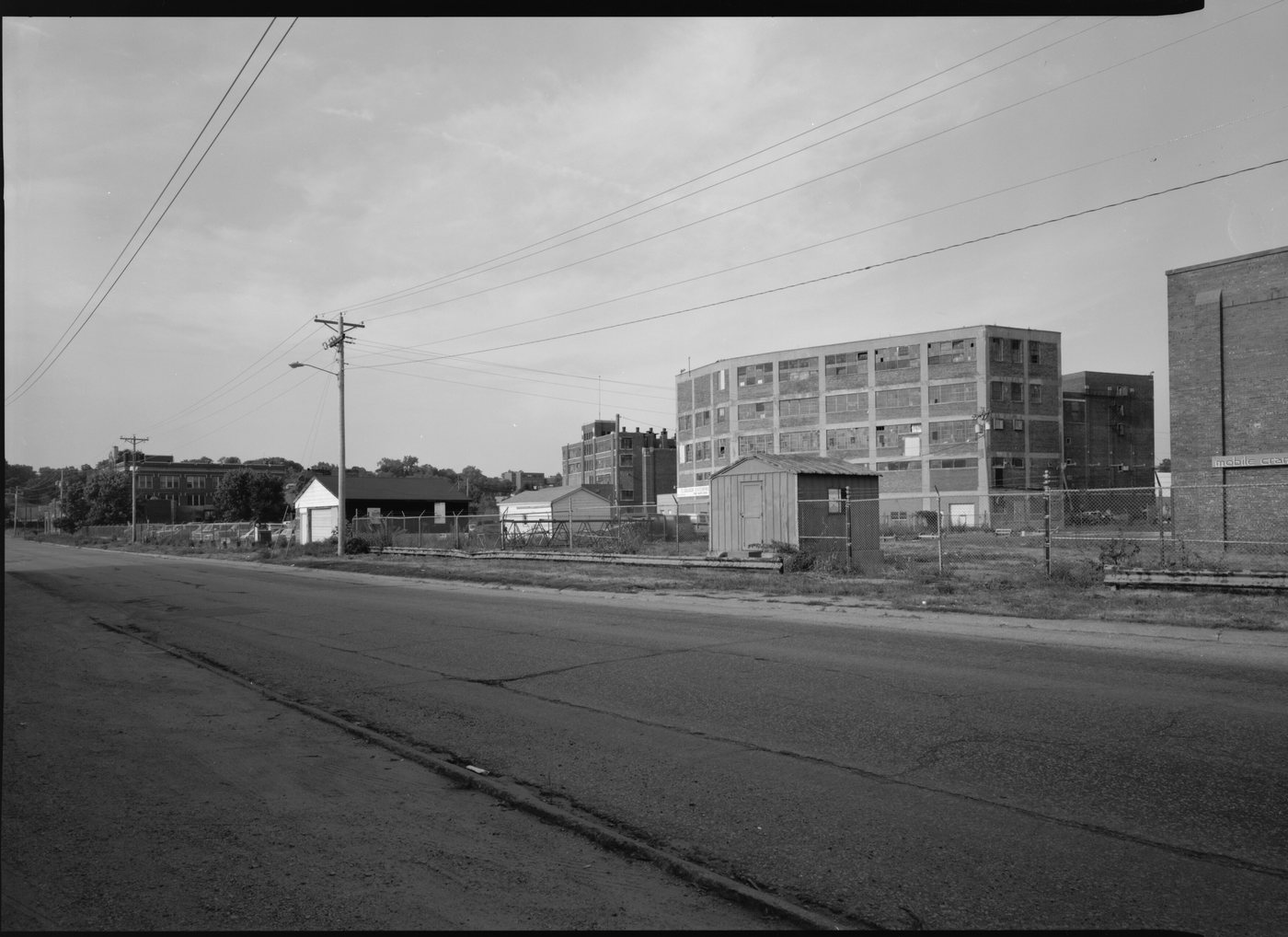 A wide street with an empty grassed lot and old brick factory buildings in the distance
