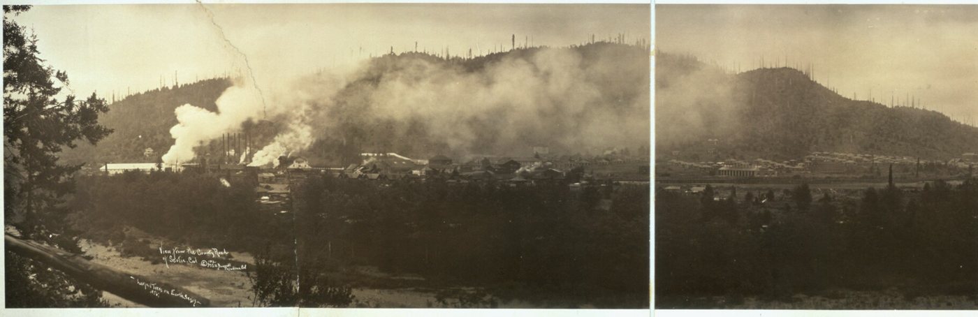 A valley seen from a road above a river: smoke pouring from mill stacks, the town spread beneath logged-over hills