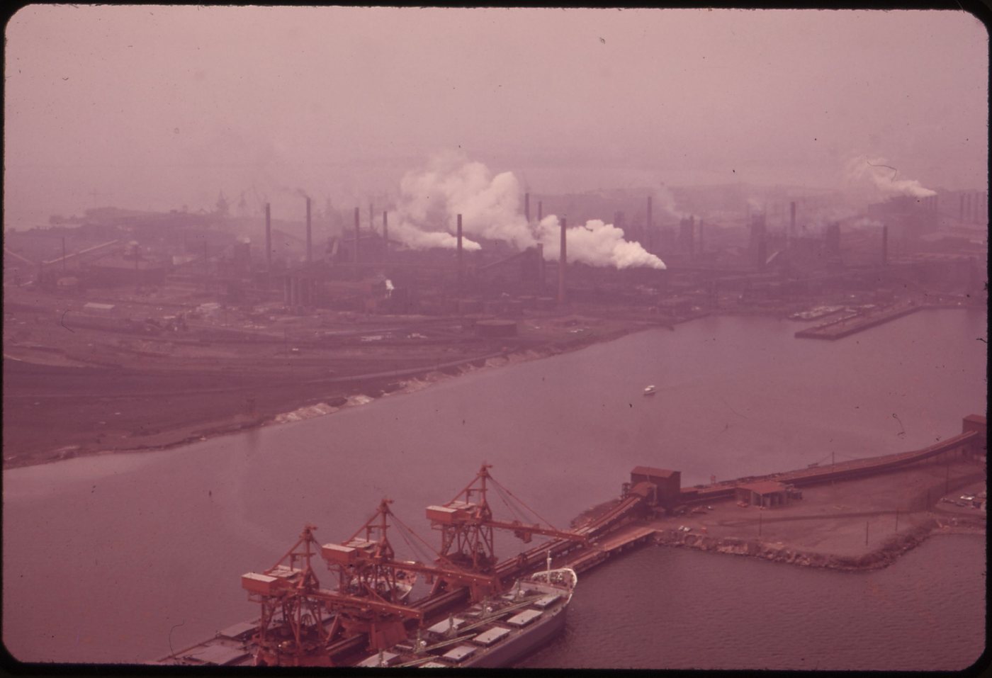 A hazy aerial view of the steelworks spread along the shore, with ore piers and cranes in the water