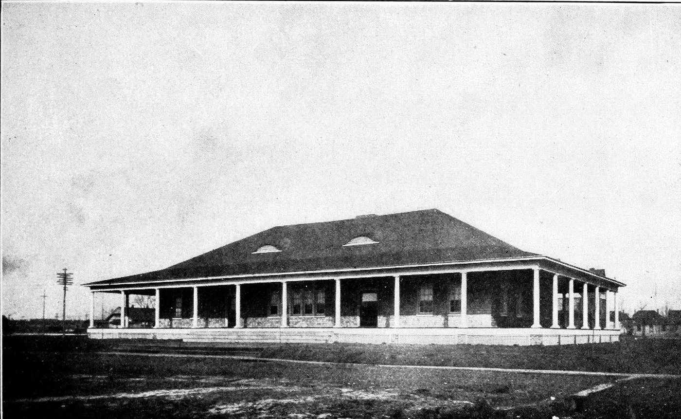 A long single-storey clubhouse with a deep hipped roof and a veranda running all the way round, on open ground
